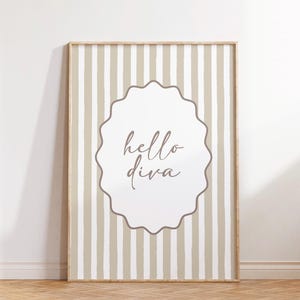 May include: A framed print with a beige and white striped background. The print features a white, scalloped-edged oval with the words "hello diva" in a cursive font. The frame is a light brown colour.