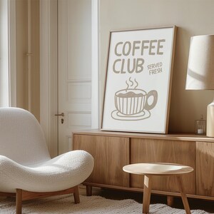 May include: A framed print with the words "COFFEE CLUB SERVED FRESH" and a coffee cup illustration in brown. The print is displayed on a wooden cabinet, with a white chair and small wooden table in the foreground. The room has a neutral color palette.