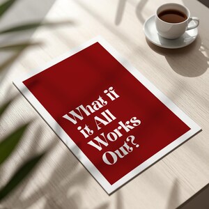 May include: A red and white print with the text "What if it All Works Out?" is displayed on a light-colored wooden surface. A white teacup and saucer with coffee are in the background.