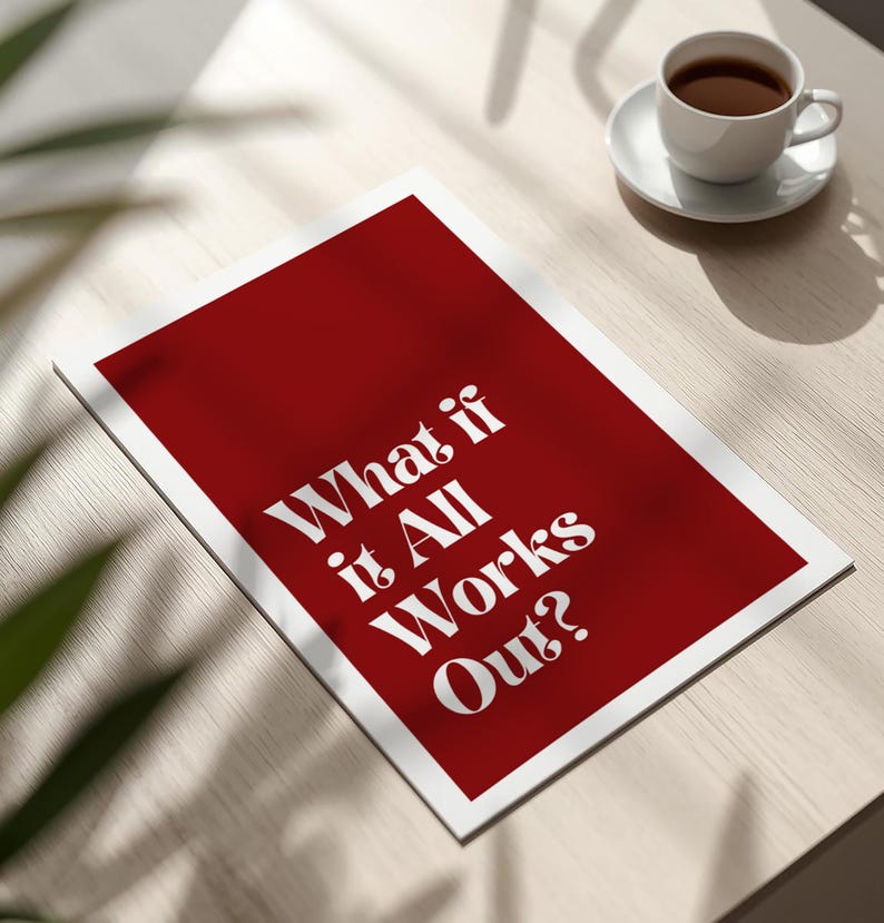 May include: A red and white print with the text "What if it All Works Out?" is displayed on a light-colored wooden surface. A white teacup and saucer with coffee are in the background.