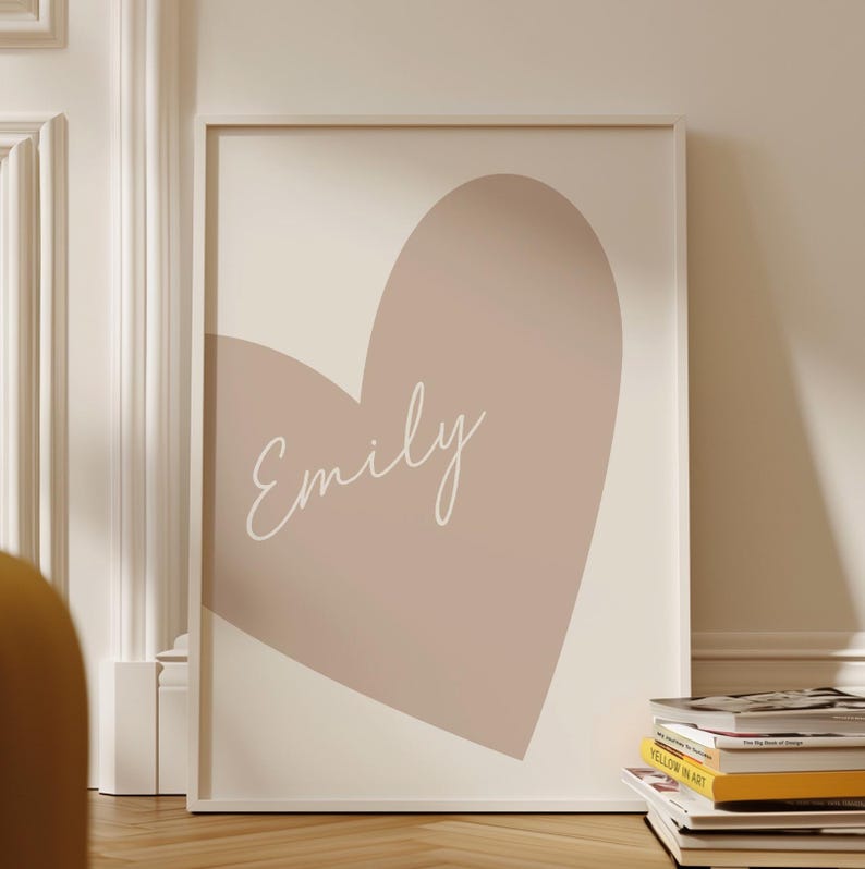 May include: Framed art print featuring a large, muted pink heart shape against a cream background. The name "Emily" is written in a delicate, cursive font across the heart. The artwork is displayed in a white frame.