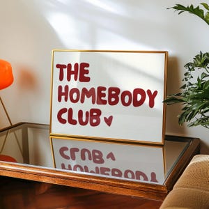 May include: A framed print with a gold frame and white background. The text "THE HOMEBODY CLUB" is written in red with a small heart symbol.
