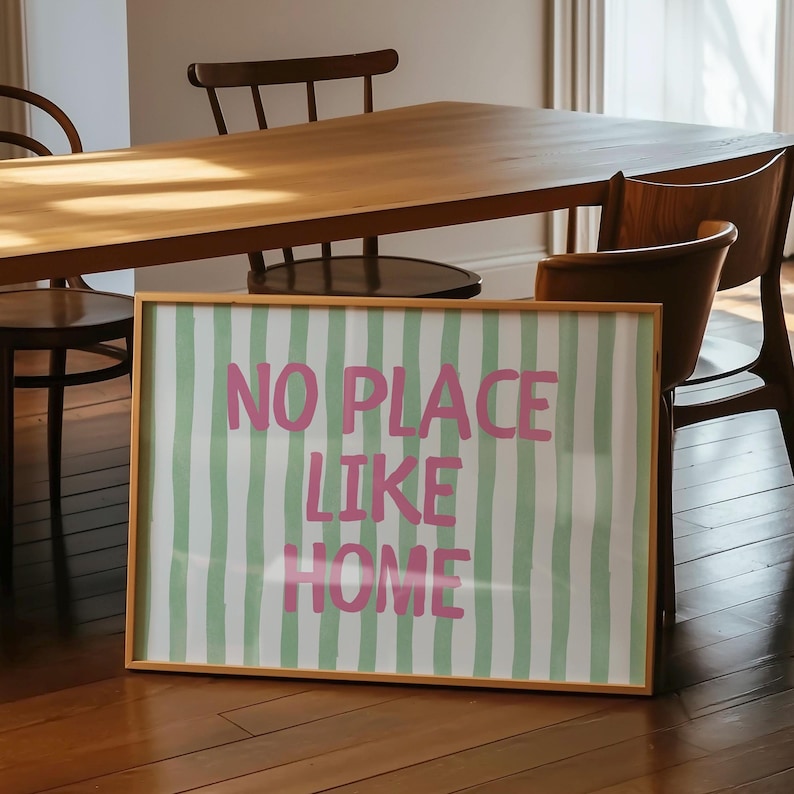 May include: A framed print with the text "NO PLACE LIKE HOME" in pink letters. The background features vertical green and white stripes. The frame is a light brown colour. The print is displayed in a room with wooden furniture.