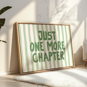 May include: A framed canvas art print with a light wood frame. The artwork features a green and white striped background with the words "JUST ONE MORE CHAPTER" in green block letters. The print is leaning against a wall.