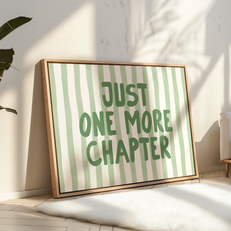 May include: A framed canvas art print with a light wood frame. The artwork features a green and white striped background with the words "JUST ONE MORE CHAPTER" in green block letters. The print is leaning against a wall.