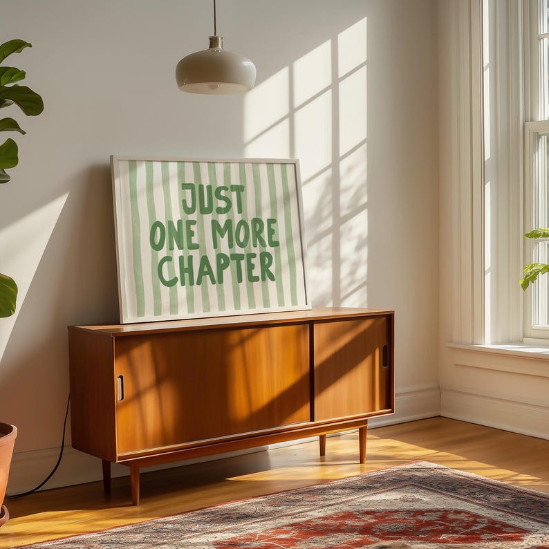 May include: A framed print with the text "JUST ONE MORE CHAPTER" in green lettering on a striped background. The print rests on a mid-century modern wooden cabinet. The cabinet has sliding doors and is set on thin legs. A rug is visible in the foreground.