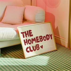 May include: A gold framed sign with the text "THE HOMEBODY CLUB" in red lettering with a small pink heart.