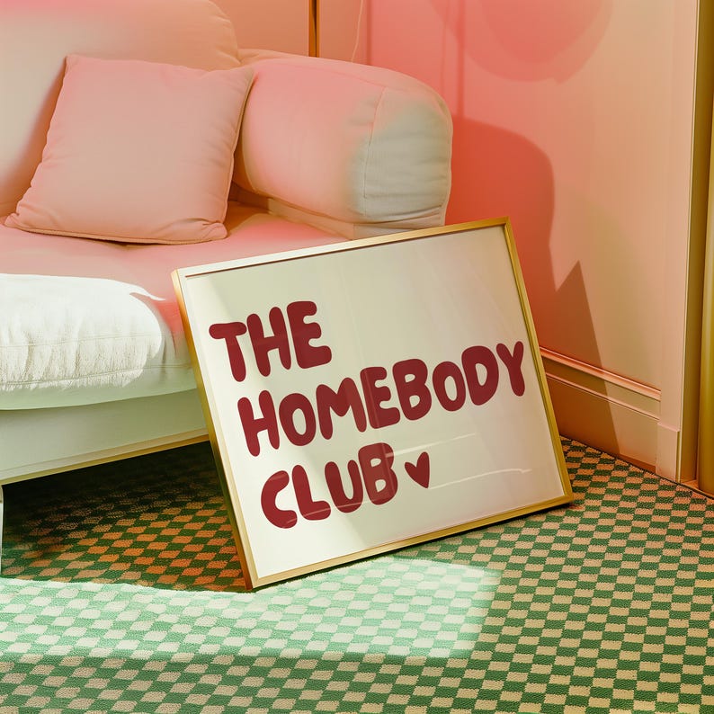 May include: A gold framed sign with the text "THE HOMEBODY CLUB" in red lettering with a small pink heart.
