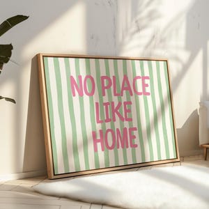 May include: A framed canvas print featuring the phrase "NO PLACE LIKE HOME" in pink lettering, set against a backdrop of green and white vertical stripes. The artwork is displayed in a room with natural light.