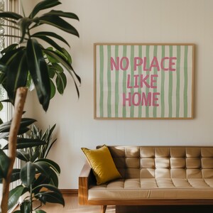 May include: A framed print with the words "NO PLACE LIKE HOME" in pink on a green and white striped background. The artwork is above a brown leather sofa with a yellow cushion. A large green plant is in the foreground.