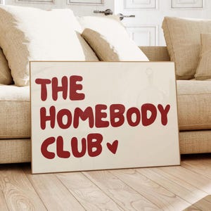 May include: A white poster with a brown frame, featuring the text "THE HOMEBODY CLUB" in red, with a small red heart symbol.
