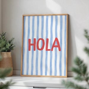 May include: A framed art print with a light wood frame. The print features a blue and white striped background with the word "HOLA" in red block letters. The artwork is displayed on a white surface.