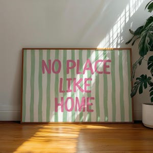 May include: A framed artwork with the text "NO PLACE LIKE HOME" in pink, set against a backdrop of green and white vertical stripes. The artwork is in a brown wooden frame and is displayed on a wooden floor.