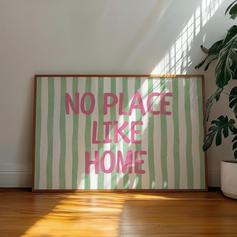 May include: A framed artwork with the text "NO PLACE LIKE HOME" in pink, set against a backdrop of green and white vertical stripes. The artwork is in a brown wooden frame and is displayed on a wooden floor.