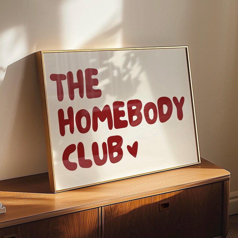 May include: A gold framed print with the text "The Homebody Club" in red lettering with a small red heart symbol. The print is hanging on a wall above a wooden dresser.