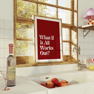 May include: A framed print with a red background and the text "What if it All Works Out?" is displayed in a kitchen setting. A bottle of wine and a glass are on the counter. Pomegranates are in the sink.