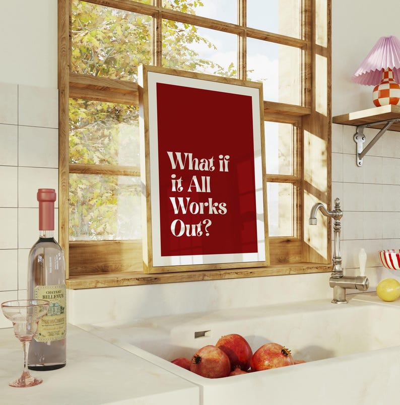 May include: A framed print with a red background and the text "What if it All Works Out?" is displayed in a kitchen setting. A bottle of wine and a glass are on the counter. Pomegranates are in the sink.