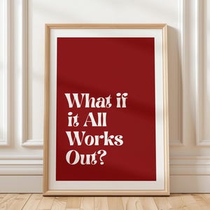 May include: A framed print with a red background and the white text "What if it All Works Out?". The print is in a light brown wooden frame and leans against a white wall with molding.