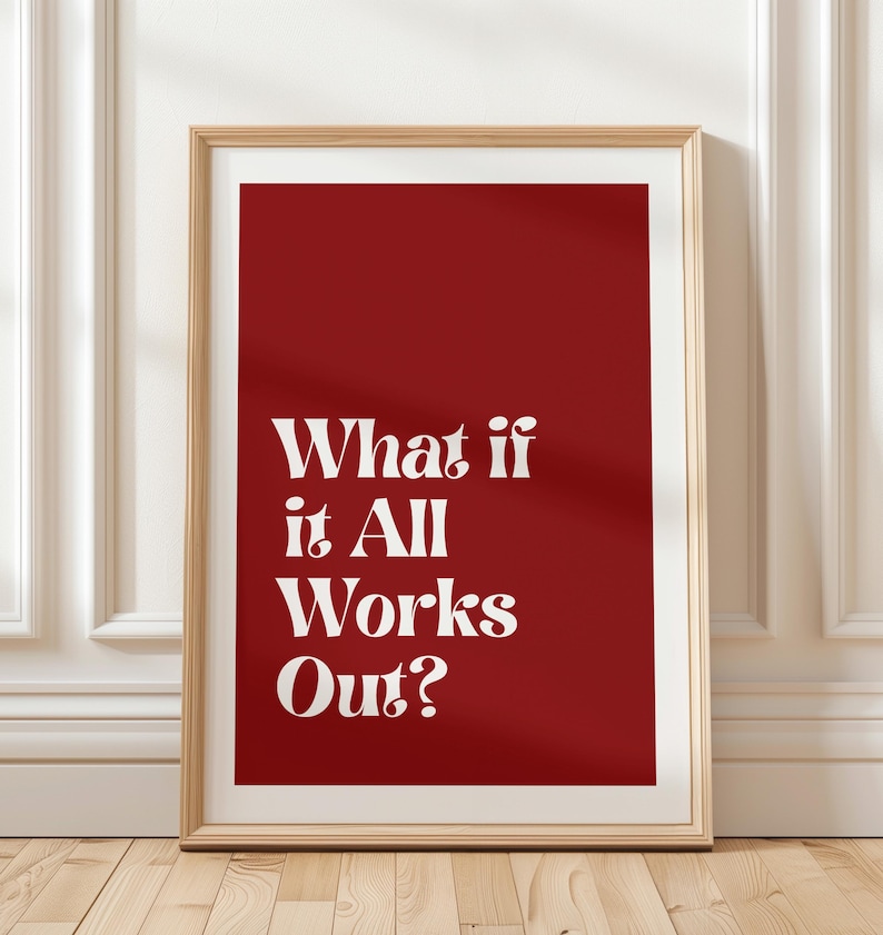 May include: A framed print with a red background and the white text "What if it All Works Out?". The print is in a light brown wooden frame and leans against a white wall with molding.