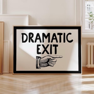 May include: A framed print featuring the words "DRAMATIC EXIT" in bold black lettering. Beneath the text is a black and white illustration of a hand pointing right. The frame is black, and the background is off-white.