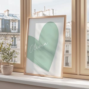 May include: A framed print featuring a large, soft green heart shape on a white background. The name "Olivia" is written in cursive across the heart. The frame is a light wood color, and the print is displayed on a windowsill.