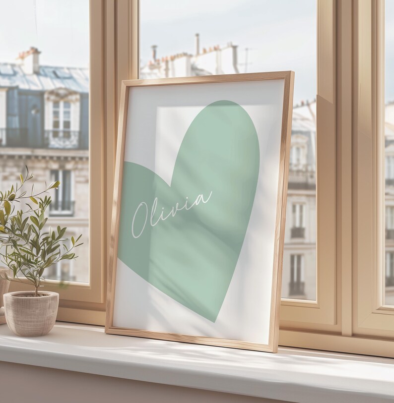 May include: A framed print featuring a large, soft green heart shape on a white background. The name "Olivia" is written in cursive across the heart. The frame is a light wood color, and the print is displayed on a windowsill.