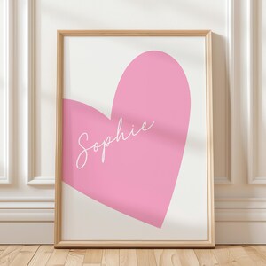 May include: A framed art print featuring a large pink heart on a white background. The name "Sophie" is written in cursive across the heart. The frame is a light wood color, and the artwork is displayed against a white wall.