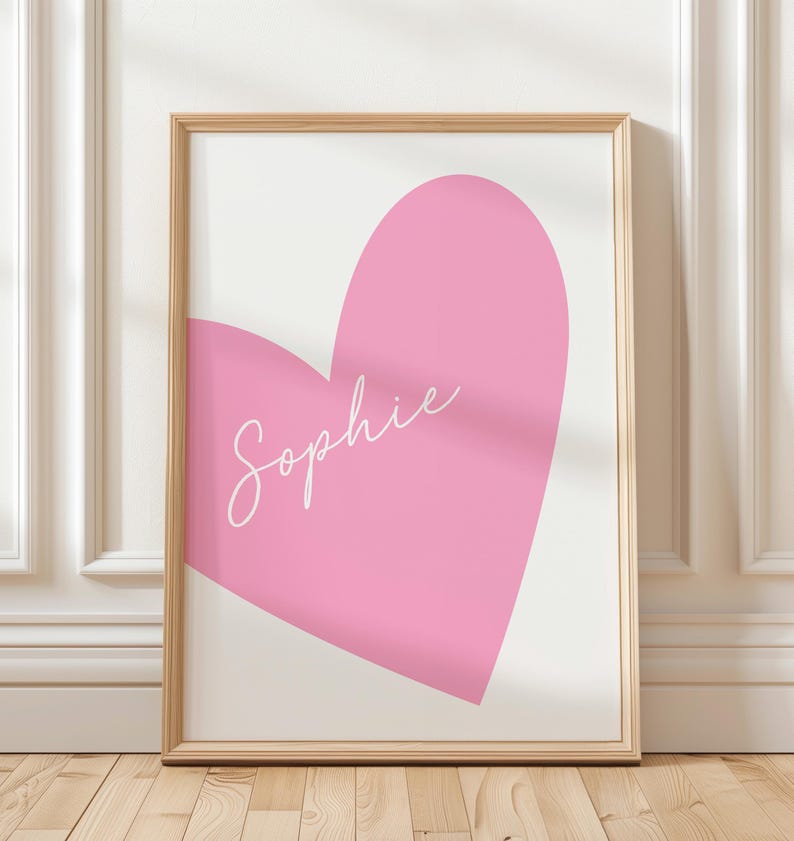 May include: A framed art print featuring a large pink heart on a white background. The name "Sophie" is written in cursive across the heart. The frame is a light wood color, and the artwork is displayed against a white wall.