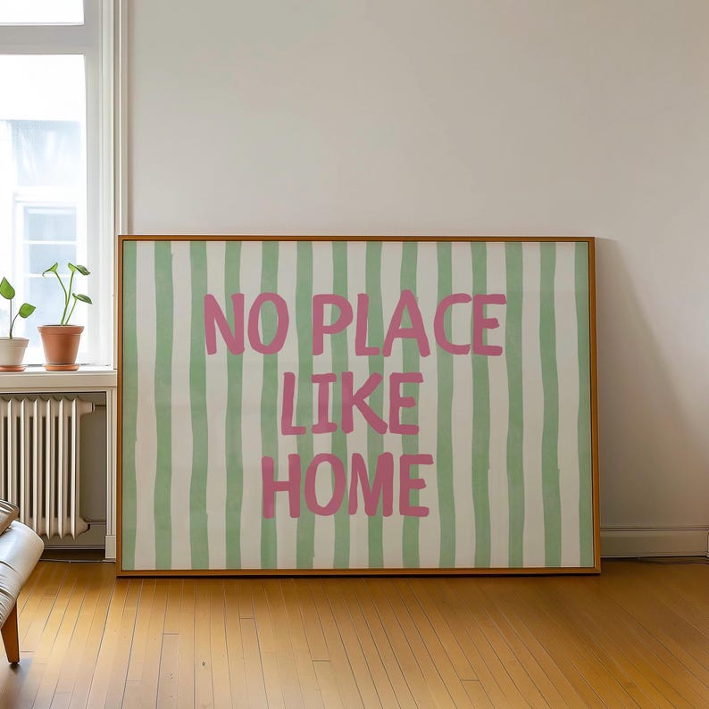 May include: A framed artwork featuring a green and white striped design with the pink text "NO PLACE LIKE HOME". The artwork is displayed in a room with wooden flooring.