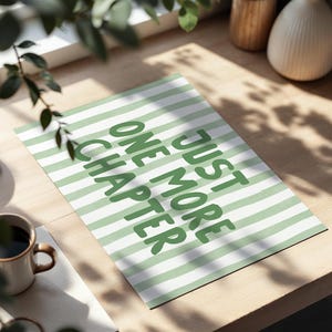 May include: A rectangular print with a green and white striped pattern. The print features the text "JUST ONE MORE CHAPTER" in green, bold letters. A coffee mug is visible in the lower left corner.