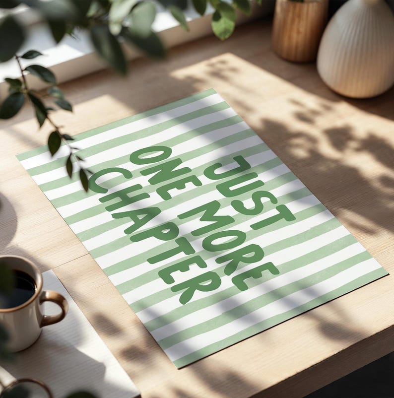 May include: A rectangular print with a green and white striped pattern. The print features the text "JUST ONE MORE CHAPTER" in green, bold letters. A coffee mug is visible in the lower left corner.