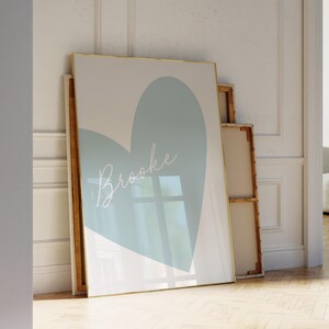 May include: A framed art print featuring a large, light blue heart shape on a white background. The name "Brooke" is written in cursive across the heart. The frame has a gold border. The artwork is leaning against a wall.