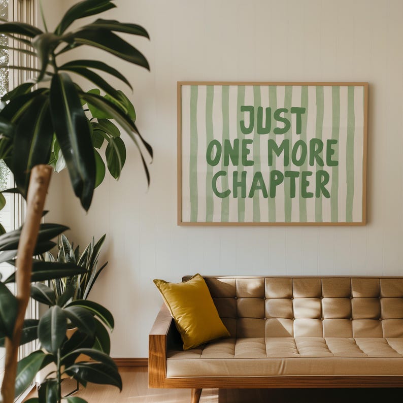 May include: A framed print with the text "JUST ONE MORE CHAPTER" in green lettering, set against a background of vertical green stripes. The print hangs above a tan leather couch with a yellow pillow. A large green plant is in the foreground.