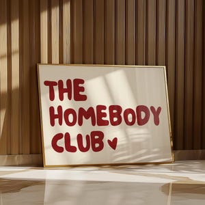 May include: A white framed print with the text "THE HOMEBODY CLUB" in red lettering with a small red heart symbol.