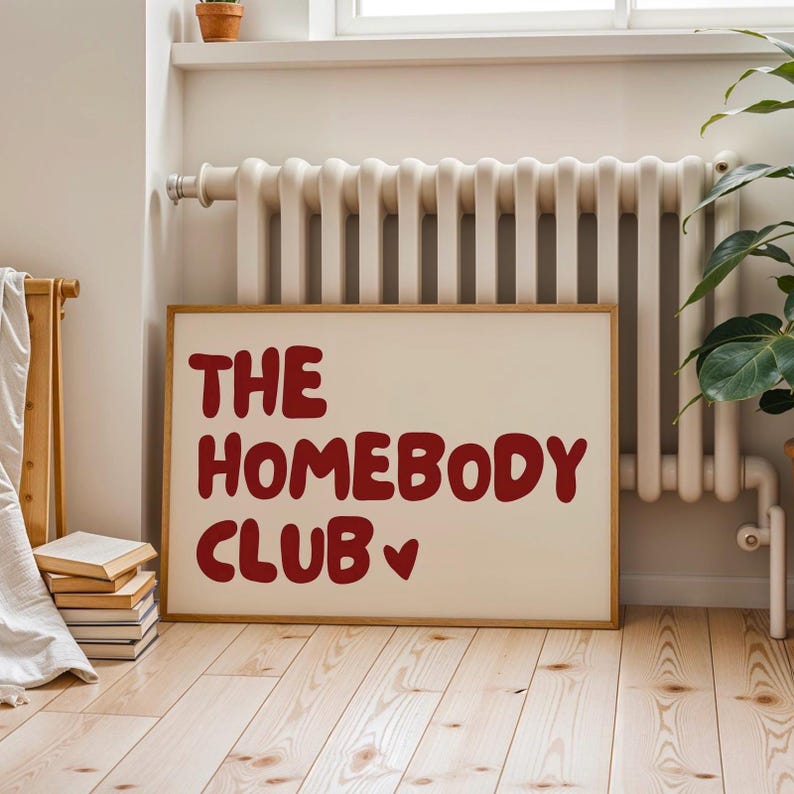 May include: A white framed poster with the text "The Homebody Club" in red lettering with a red heart symbol. The poster is hanging on a wall with a white radiator behind it.