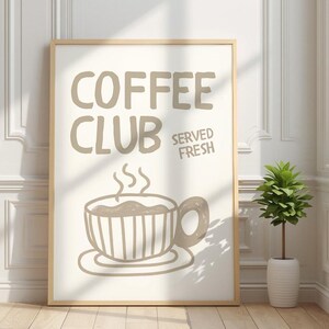 May include: A framed print with the words "COFFEE CLUB SERVED FRESH" in taupe lettering. Below the text is a drawing of a steaming coffee cup. The print is in a light wood frame and is displayed against a white wall.