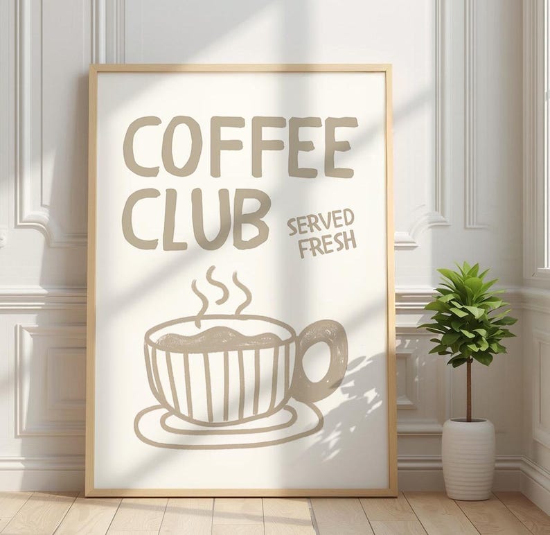 May include: A framed print with the words "COFFEE CLUB SERVED FRESH" in taupe lettering. Below the text is a drawing of a steaming coffee cup. The print is in a light wood frame and is displayed against a white wall.