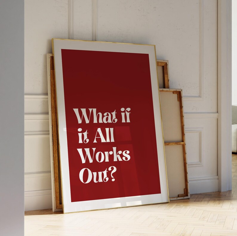 May include: A framed art print with a gold border and a deep red background. The white text reads "What if it All Works Out?" The print leans against other framed canvases, creating a layered display. The artwork is displayed on a wooden floor.
