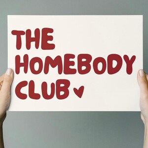 May include: A white sign with red lettering that reads "THE HOMEBODY CLUB" with a small red heart.