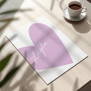 May include: A white print featuring a large lavender heart with the name "Layla" in cursive script. The print is on a wooden surface, with a cup of coffee on a saucer in the background.