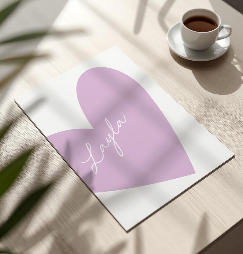 May include: A white print featuring a large lavender heart with the name "Layla" in cursive script. The print is on a wooden surface, with a cup of coffee on a saucer in the background.