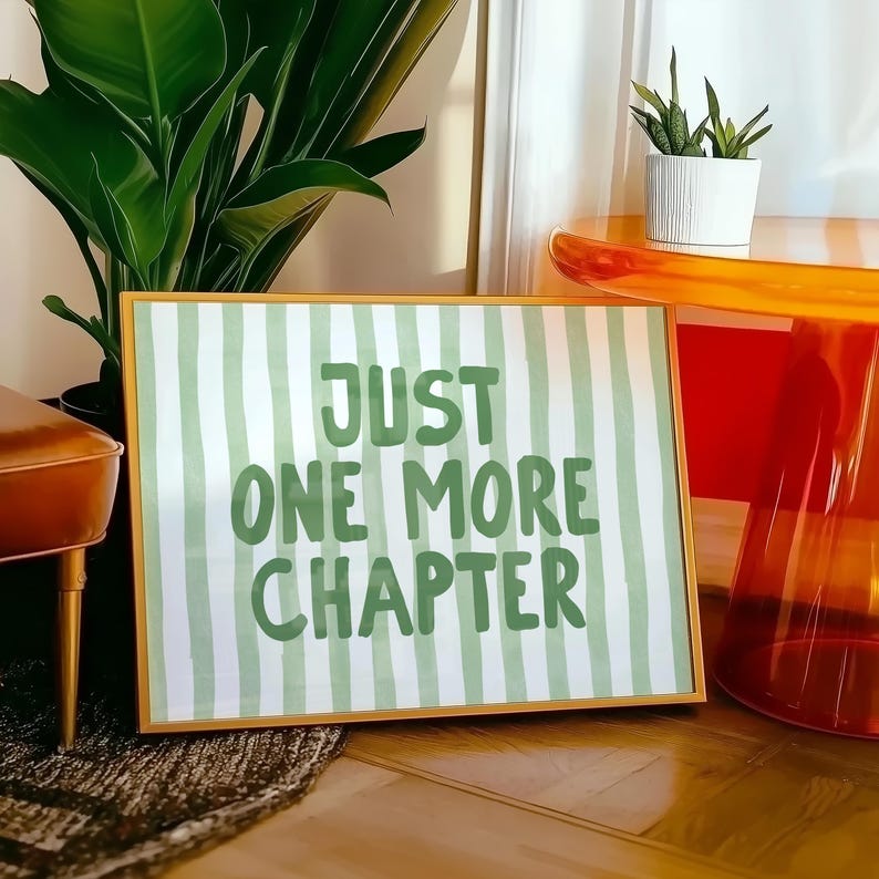 May include: A framed print with a gold border featuring the text "JUST ONE MORE CHAPTER" in green lettering on a white and green striped background. The print is displayed on a wooden surface, with plants and a modern orange table in the background.