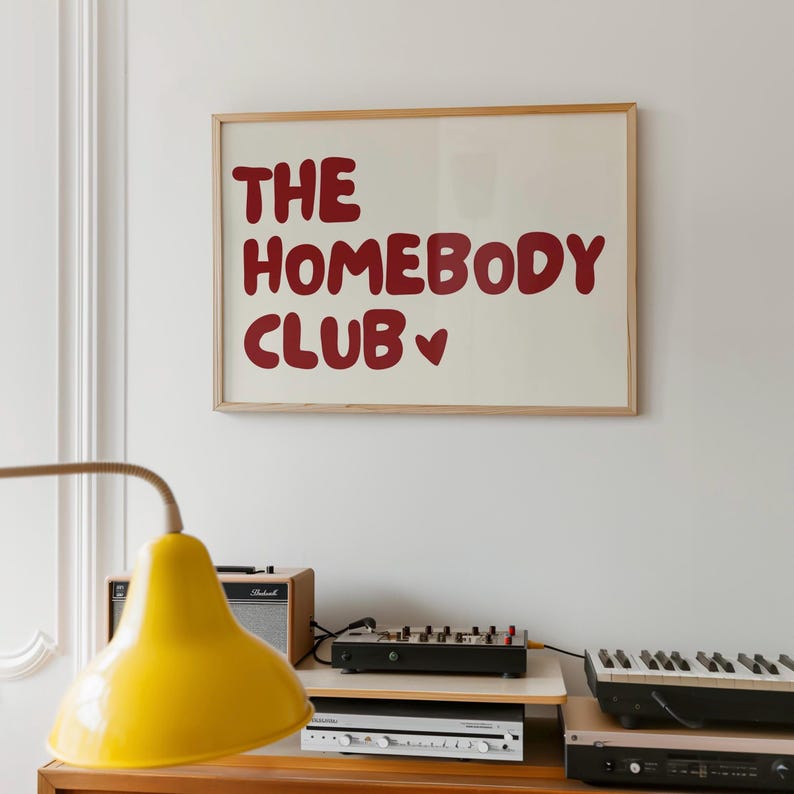 May include: A framed print with the text "The Homebody Club" in red lettering with a small heart symbol. The print is hanging on a white wall above a wooden shelf with a yellow lamp, a stereo, and a keyboard.