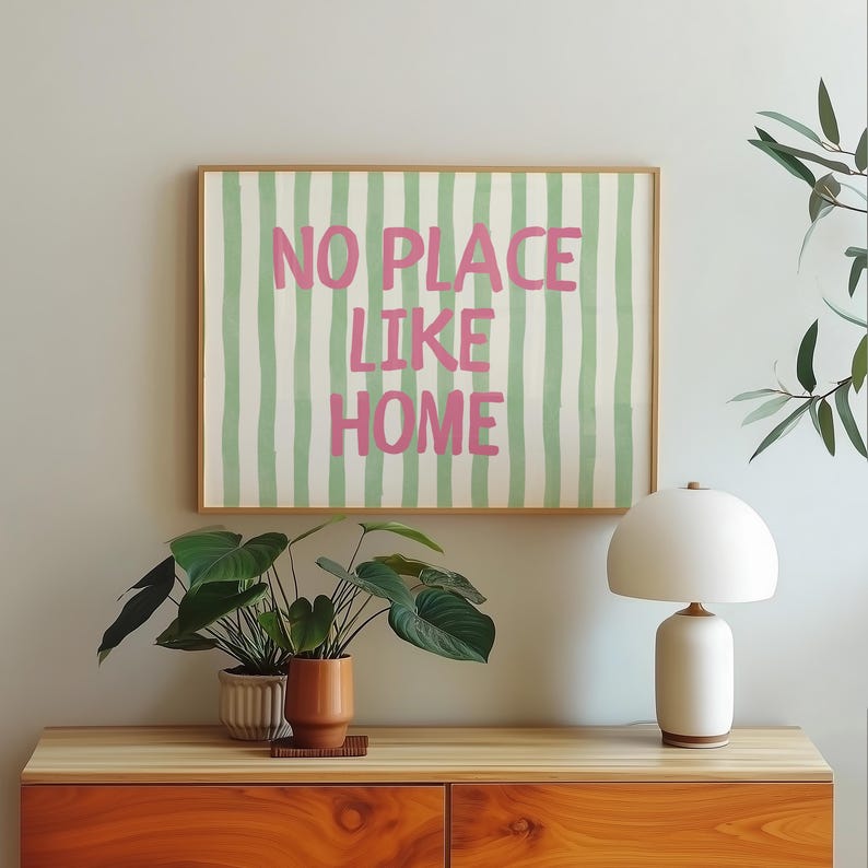 May include: A framed print with the words "NO PLACE LIKE HOME" in pink on a striped green and white background. The artwork is above a wooden cabinet, a white lamp, and potted plants.