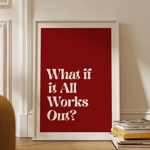 May include: A framed art print with a bold red background and the white text "What if it All Works Out?" The print is in a white frame and is leaning against a white wall. A stack of books is visible at the bottom.