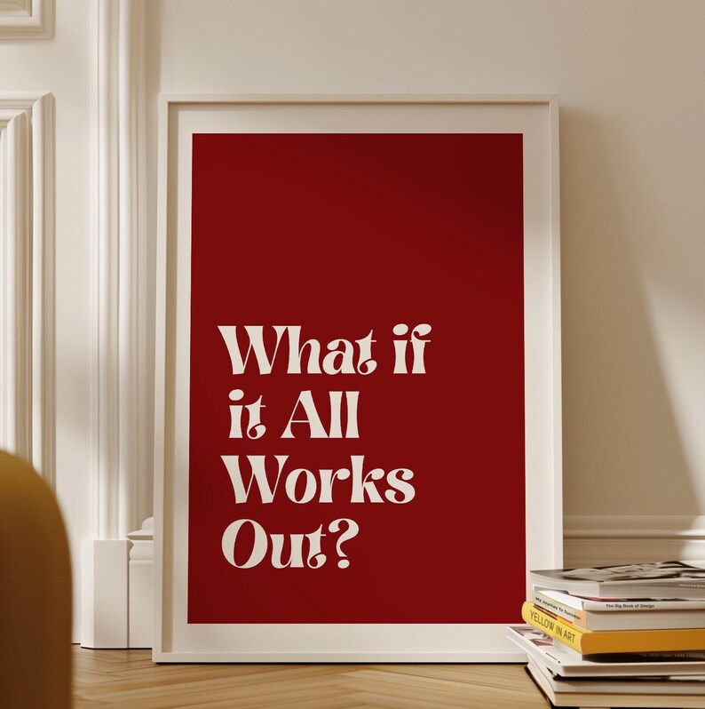 May include: A framed art print with a bold red background and the white text "What if it All Works Out?" The print is in a white frame and is leaning against a white wall. A stack of books is visible at the bottom.