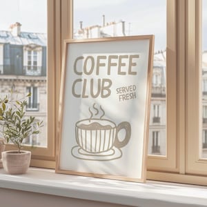 May include: A framed print with the words "COFFEE CLUB SERVED FRESH" and a coffee cup illustration. The print is in a light wood frame and sits on a windowsill with a small potted plant. The background shows a cityscape.