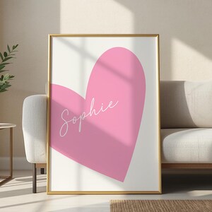 May include: A framed art print featuring a large pink heart on a white background. The name "Sophie" is written in cursive across the heart. The frame is gold.