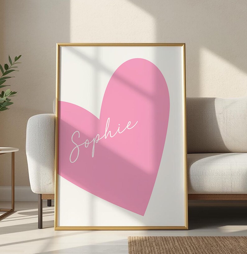 May include: A framed art print featuring a large pink heart on a white background. The name "Sophie" is written in cursive across the heart. The frame is gold.