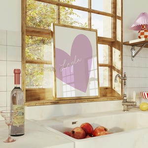 May include: A framed print featuring a large lavender heart with the name "Layla" in cursive script. The artwork is displayed on a windowsill, alongside a bottle of wine, a glass, and pomegranates.